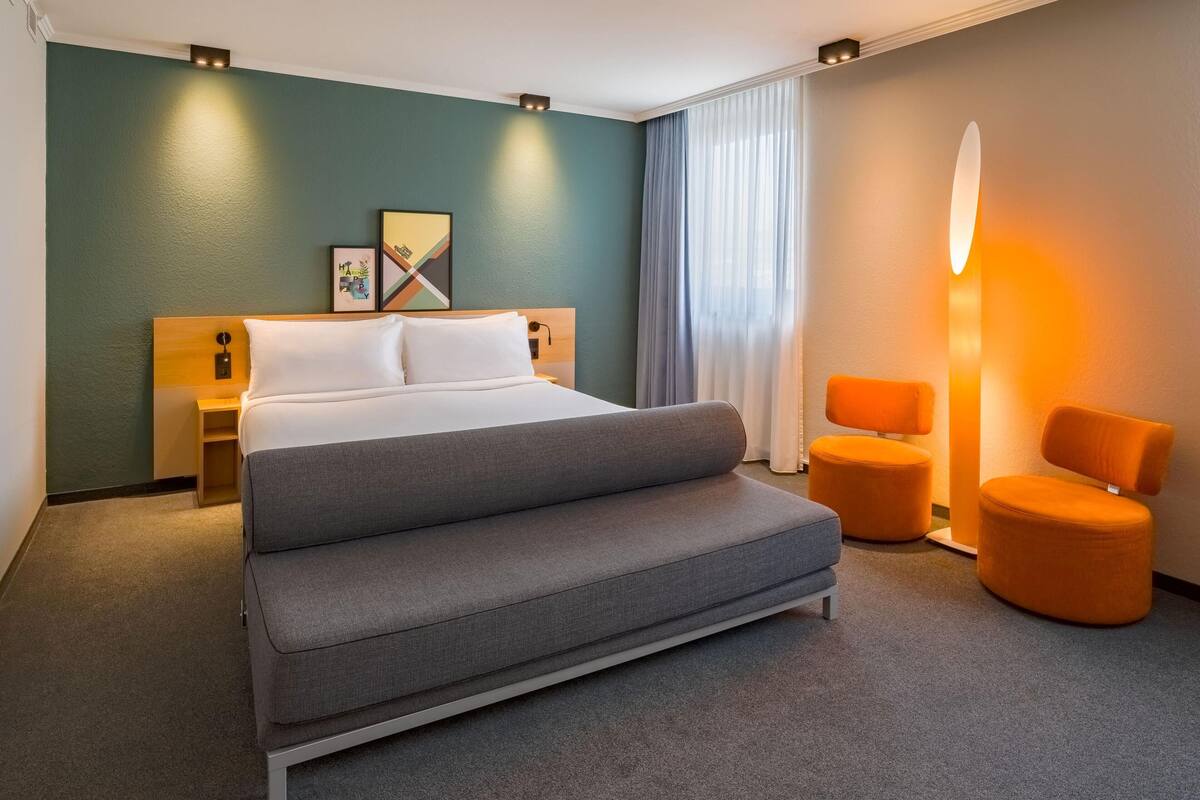 A modern hotel room with a large bed, a sofa, two orange chairs, a nightstand, and a wall-mounted lamp.