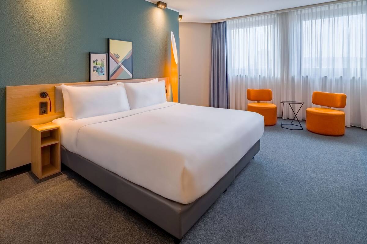 A hotel room with a large bed, a bedside table, a small side table, two orange chairs, and a large window with sheer curtains.