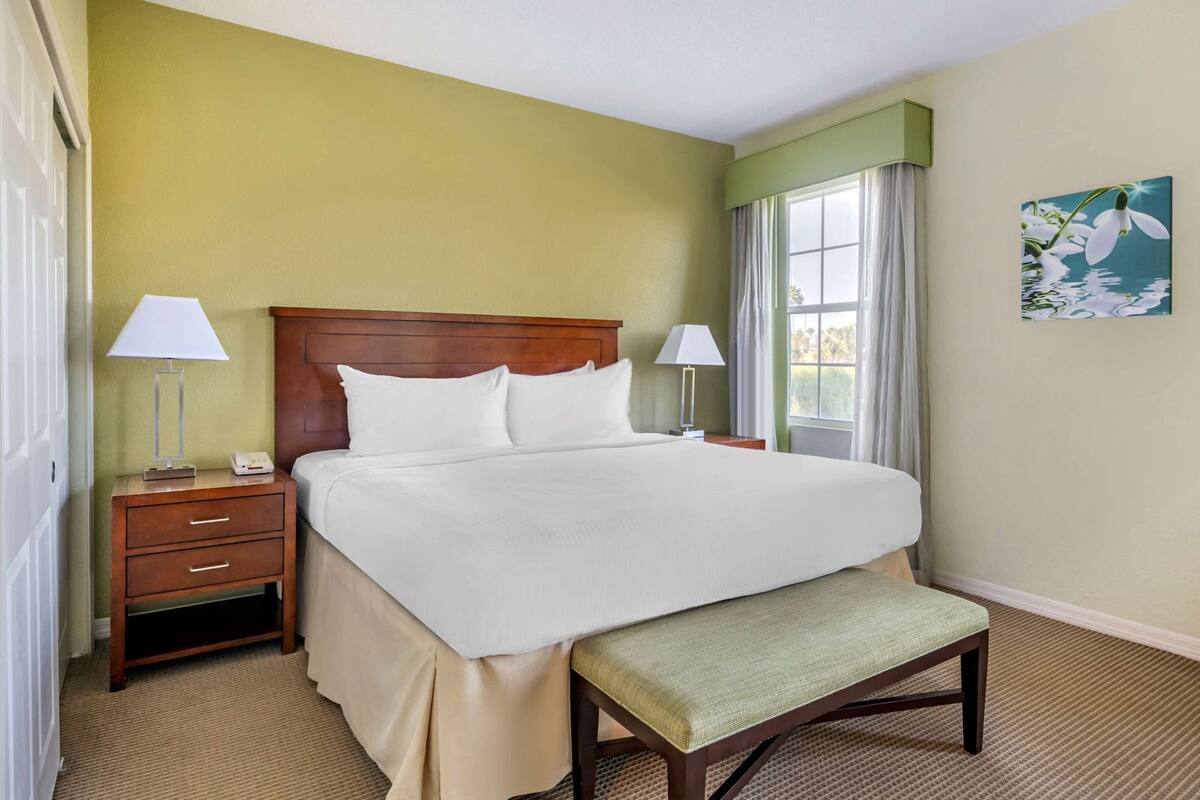 A hotel room with a large bed, two bedside tables with lamps, a bench at the foot of the bed, and a painting on the wall.