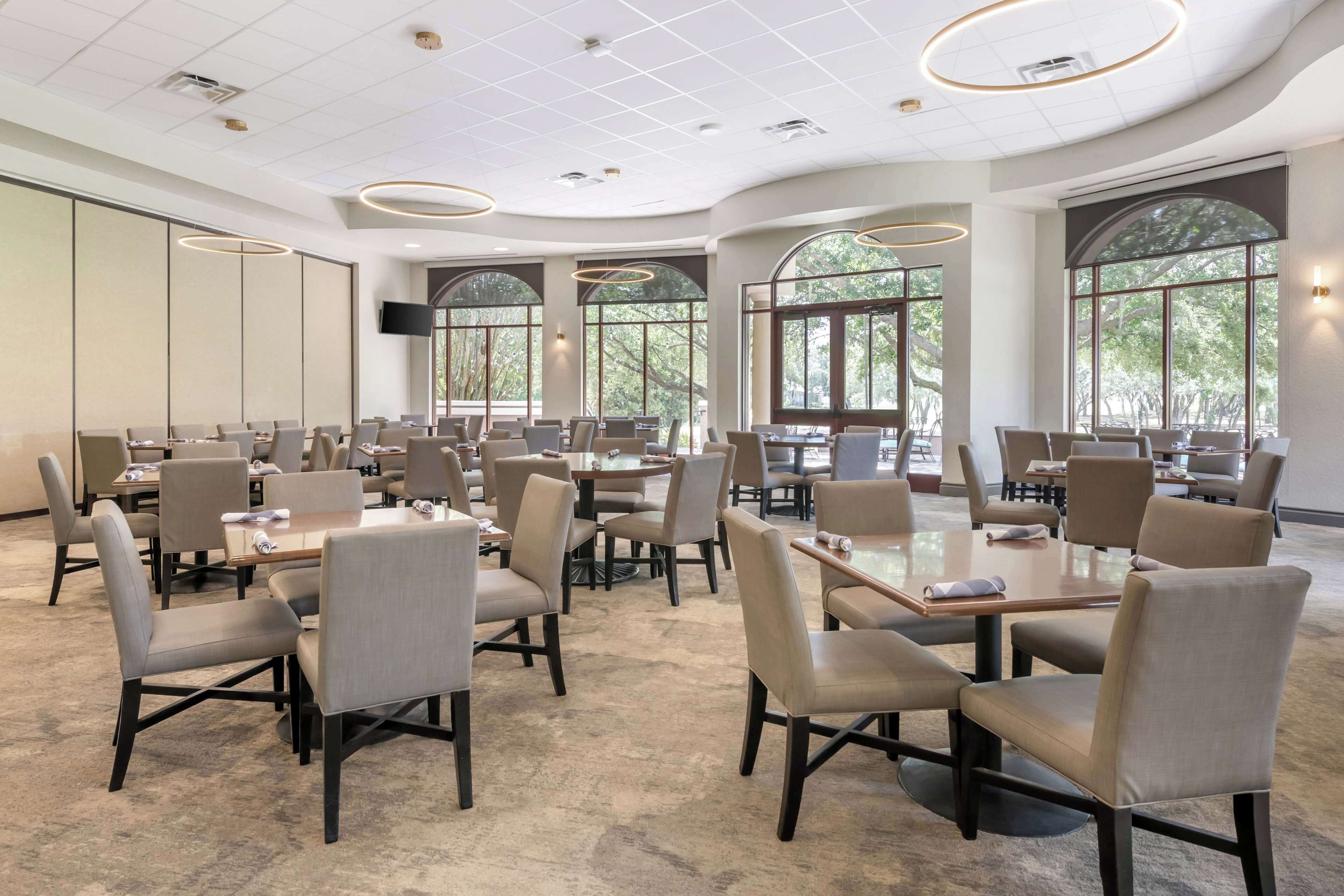 A spacious conference room with multiple tables and chairs, large windows with a view of trees, and a modern ceiling design.