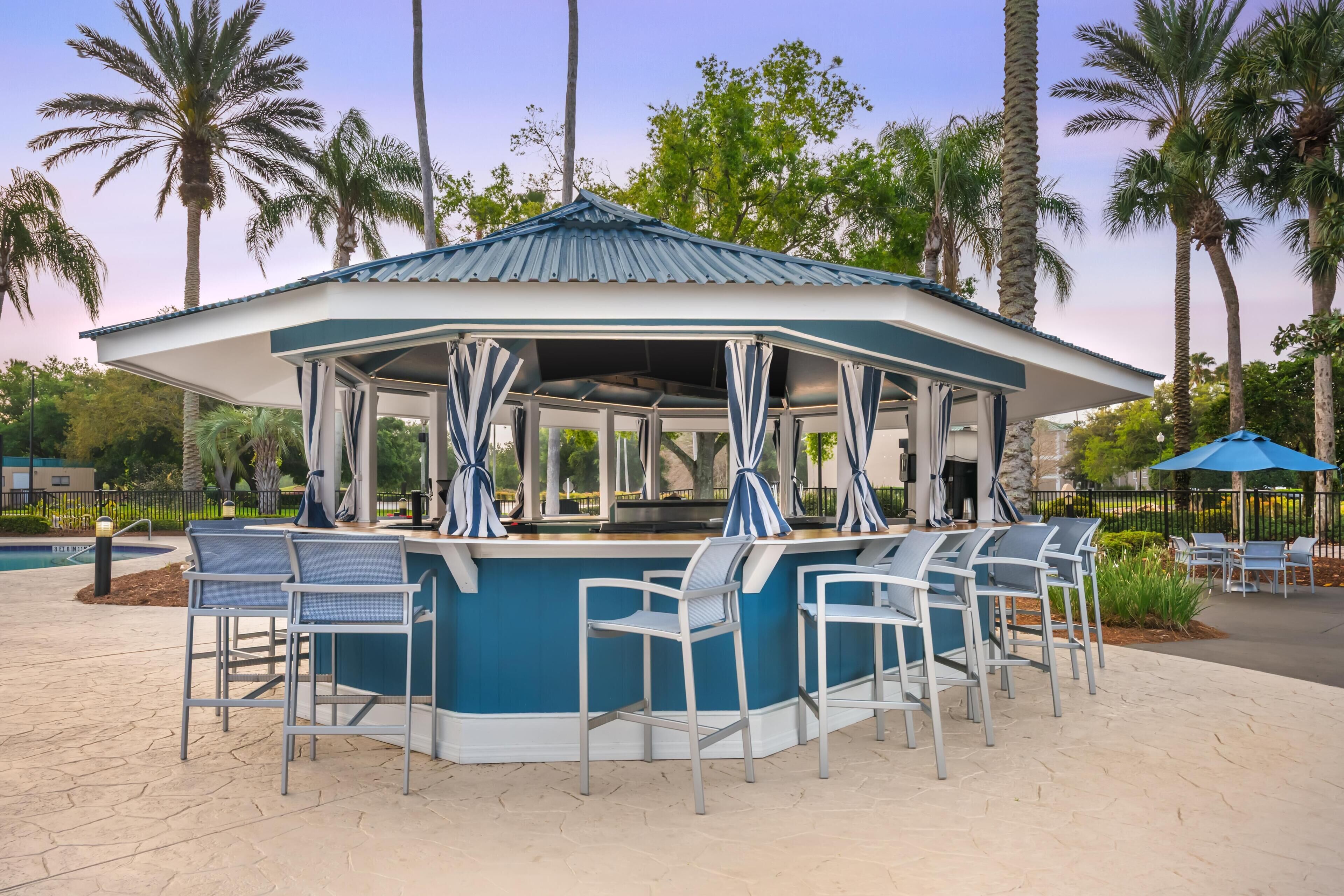 Un pabellón con decoración azul y blanca, rodeado de asientos de bar al aire libre y palmeras.