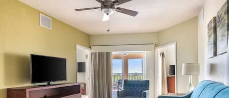 Una sala de estar con televisor de pantalla plana, mueble de madera, sofá azul, otoman y ventilador de techo.