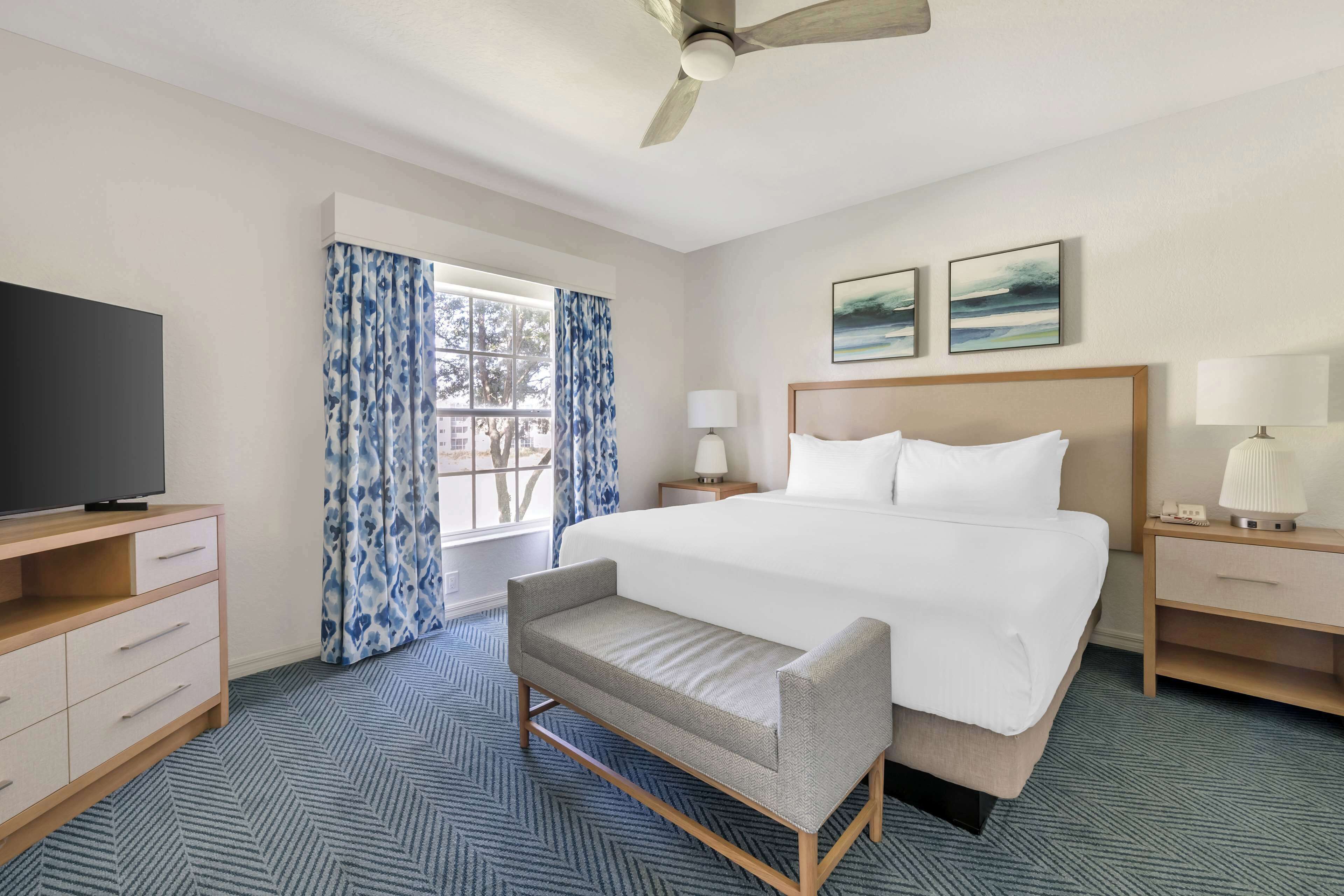 Habitación de hotel con cama, televisión, escritorio, silla y ventana con cortinas.