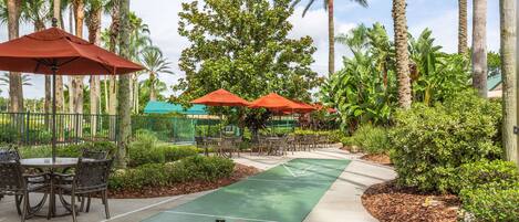 A shuffleboard court with tables and chairs, surrounded by palm trees and greenery.