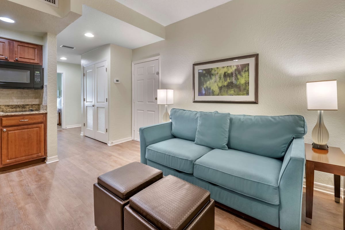 A living room with a light blue sofa, a coffee table, and a lamp. There is a kitchen area with wooden cabinets and a microwave.