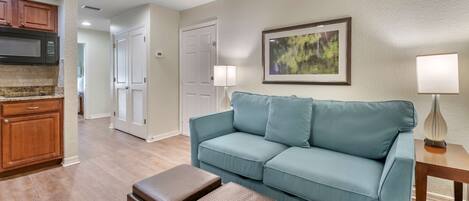 A living room with a light blue sofa, a coffee table, and a lamp. There is a kitchen area with wooden cabinets and a microwave.