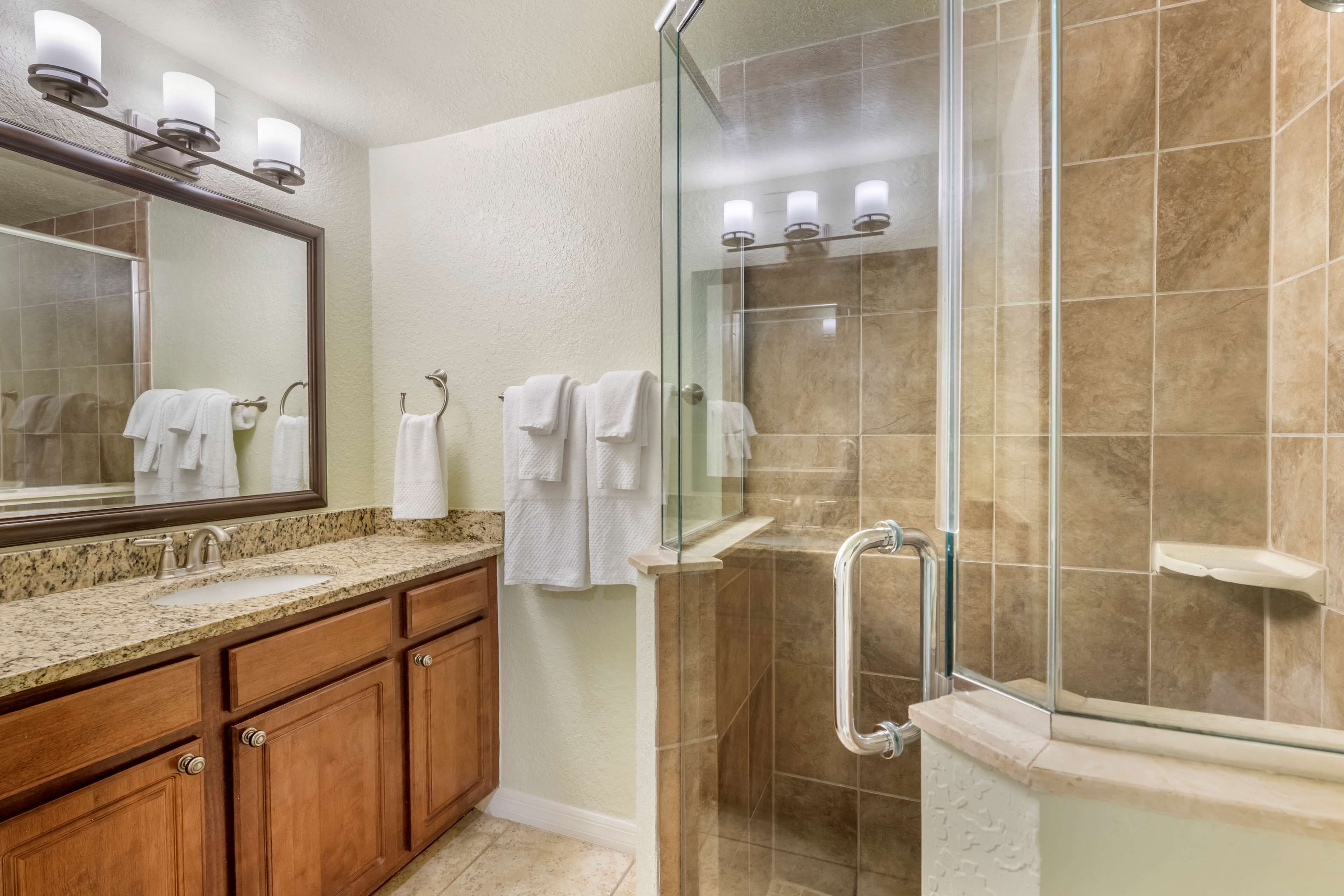 Une salle de bain dotée d’une douche à parois de verre, d’un lavabo avec un plan de travail en granit, de meubles en bois et d’un miroir avec éclairage intégré.
