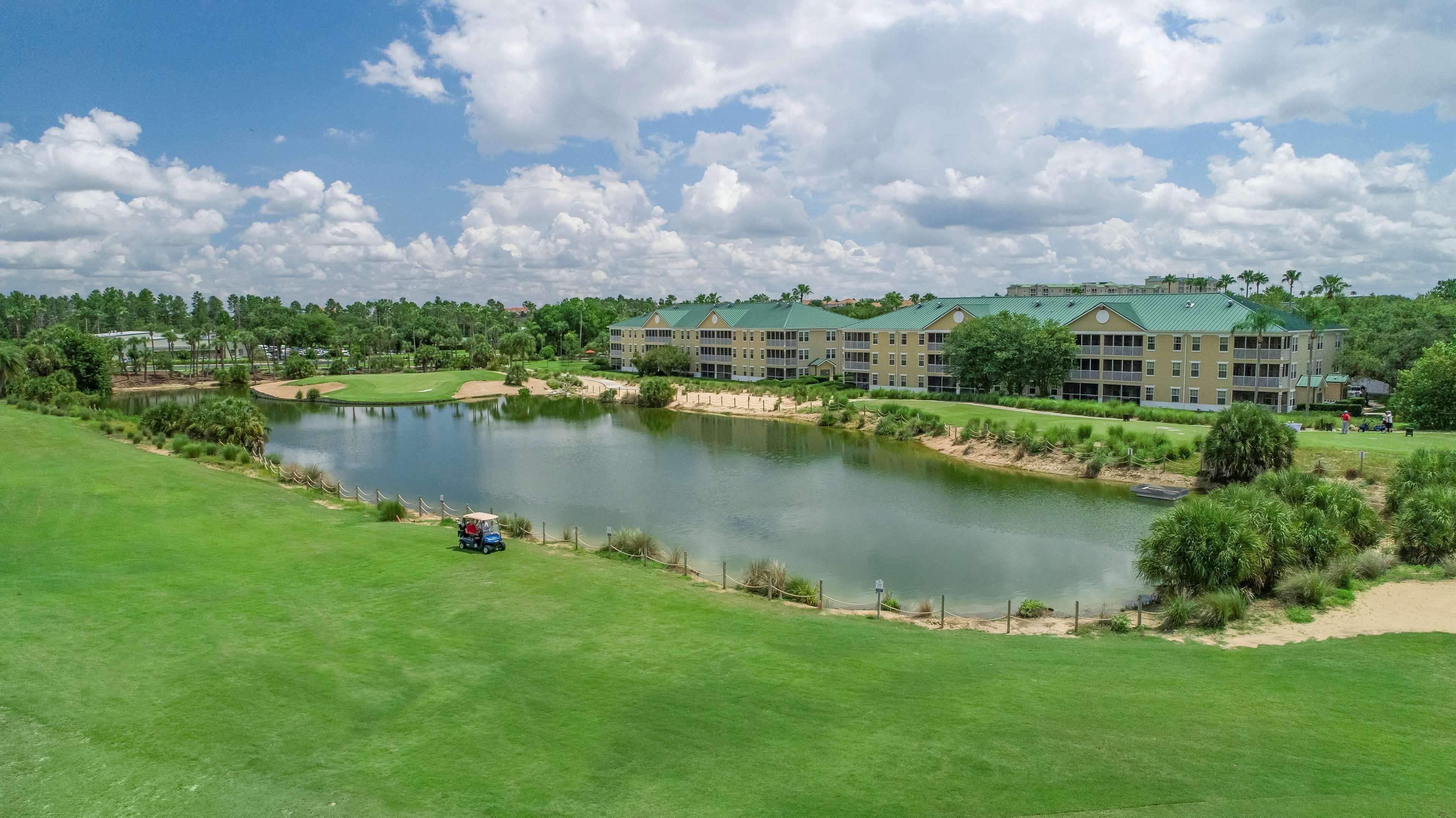 A golf course with a pond, a clubhouse, and a large building with a green roof.