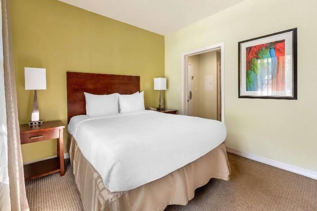 A hotel room with a large bed, two bedside tables with lamps, a door, and a framed picture on the wall.