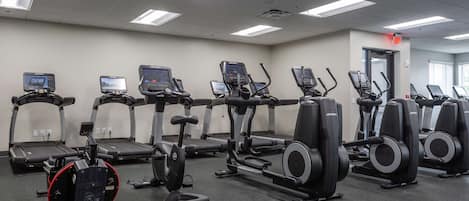 A gym with multiple treadmills and elliptical machines, a ceiling with fluorescent lights, and a door with a red emergency exit sign.