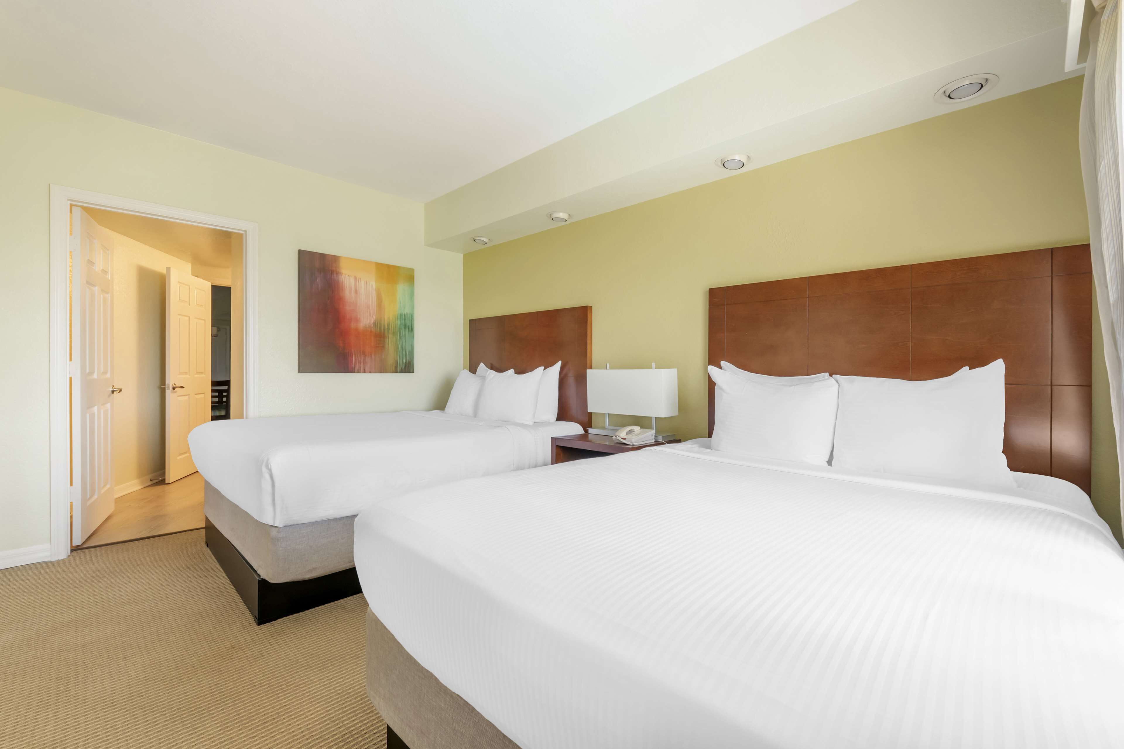 A hotel room with two beds, a bedside table with a lamp, a painting on the wall, and a door leading to another room.