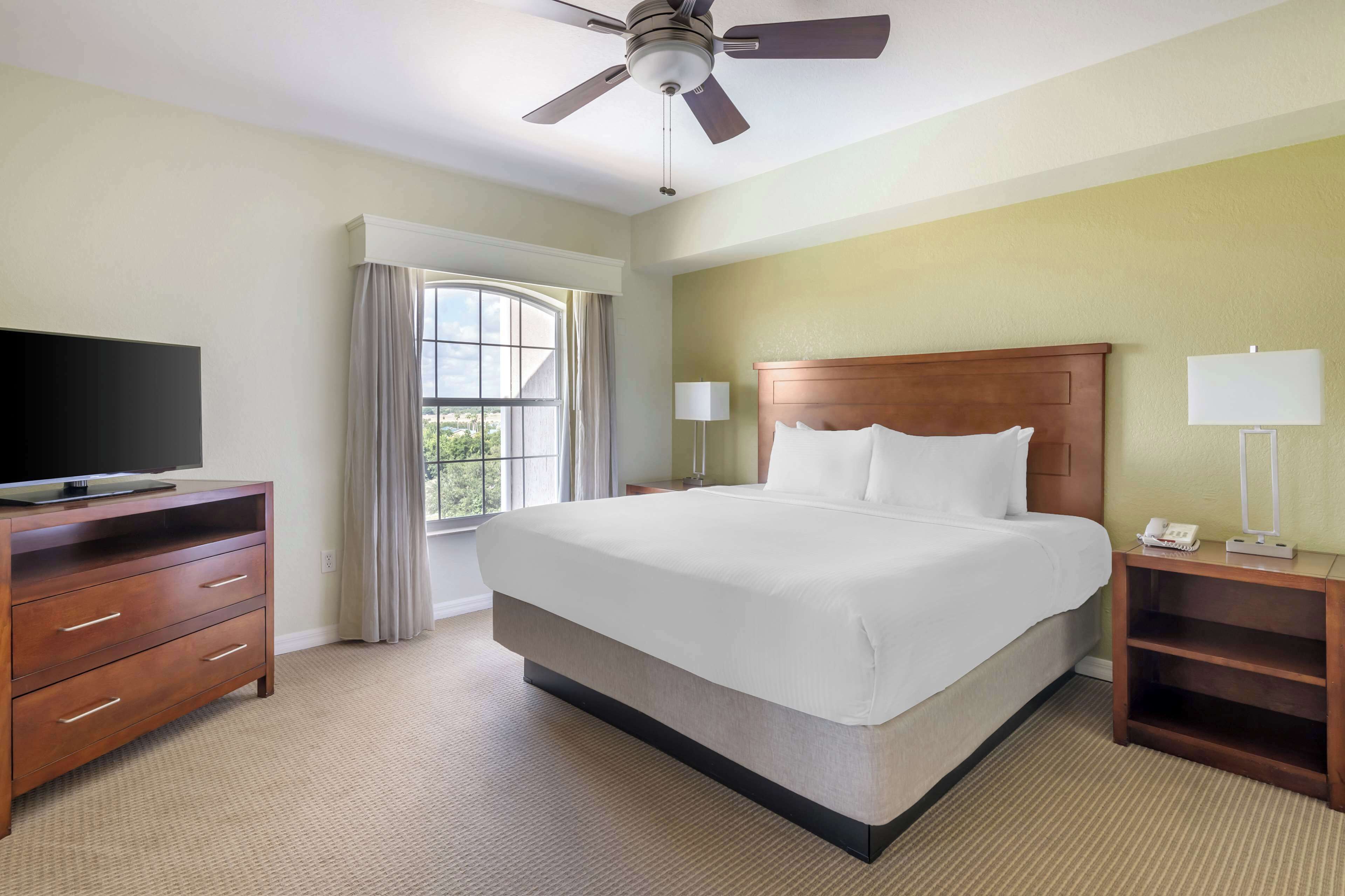 Ein Hotelzimmer mit einem großen Bett, einem Fernseher auf einer Holzkommode, einem Deckenventilator und einem Fenster mit Vorhängen.