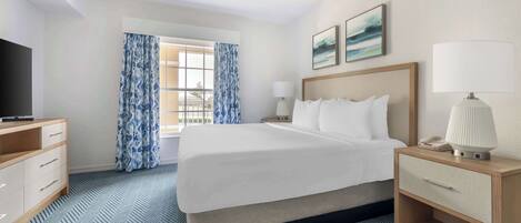 Habitación de hotel con cama, escritorio, televisión, lámpara y ventana con cortinas.