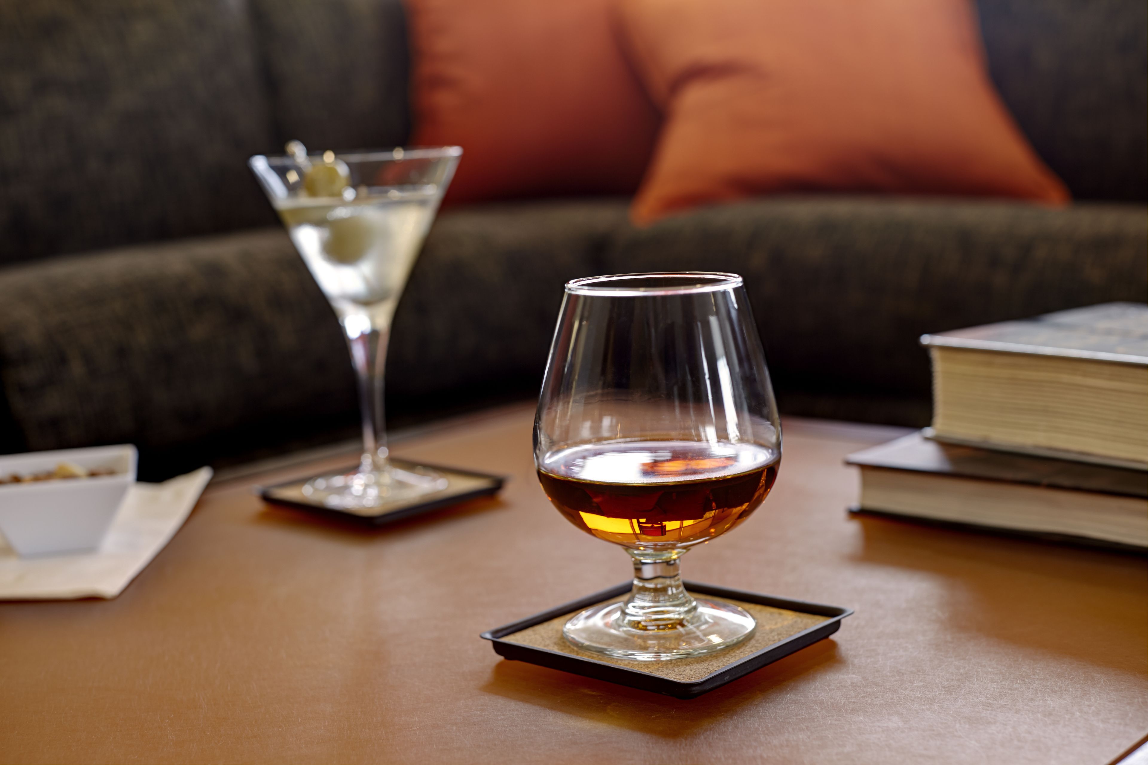 A glass of amber liquid on a coaster, a martini glass with a garnish, and a book on a table.