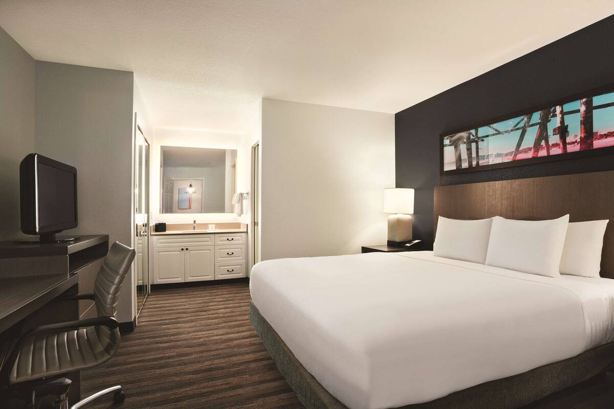 A hotel room with a large bed, a desk with a chair, a television, a bathroom with a mirror, and a painting on the wall.