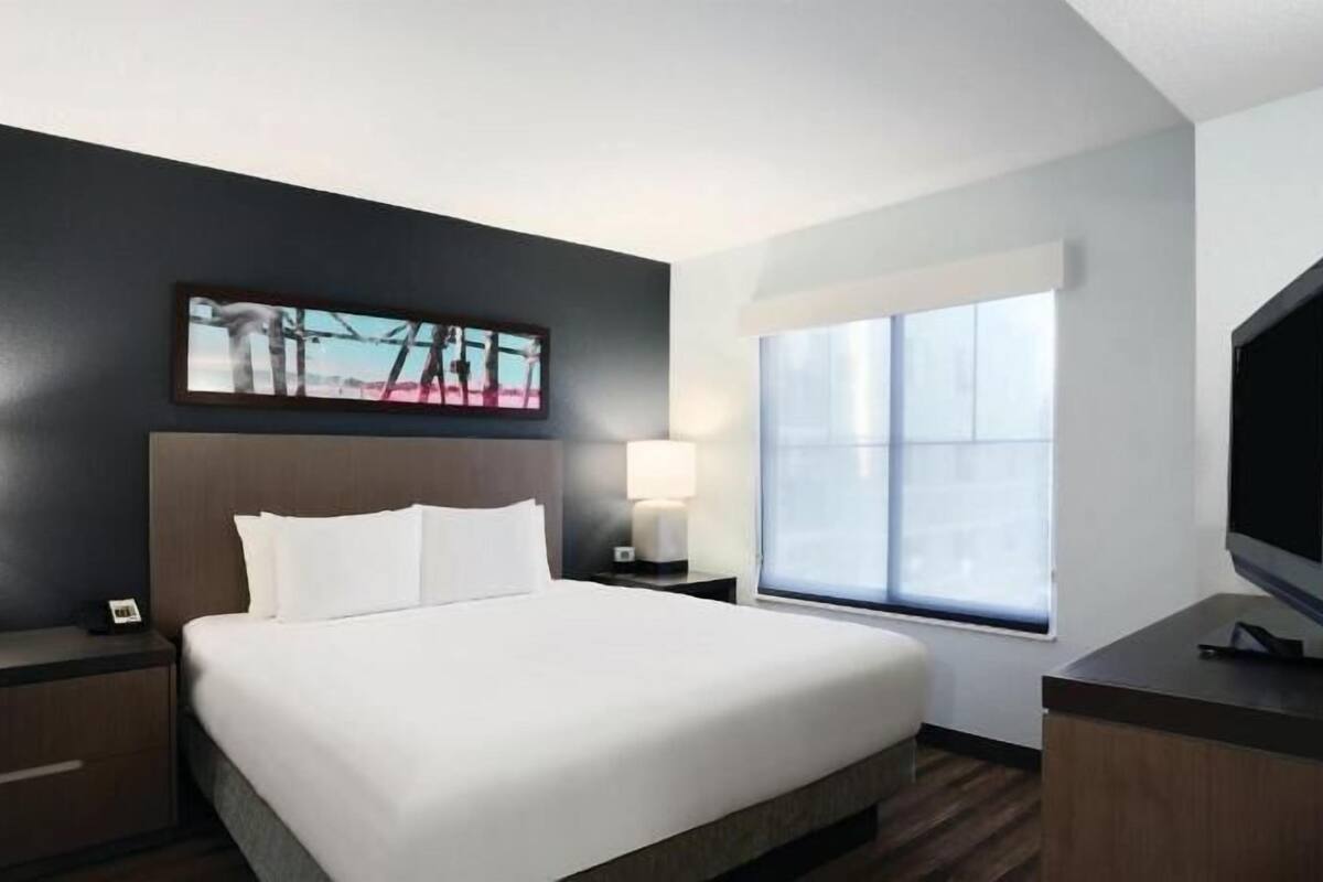 A hotel room with a bed, two bedside lamps, a flat-screen TV, a window, and a painting on the wall.