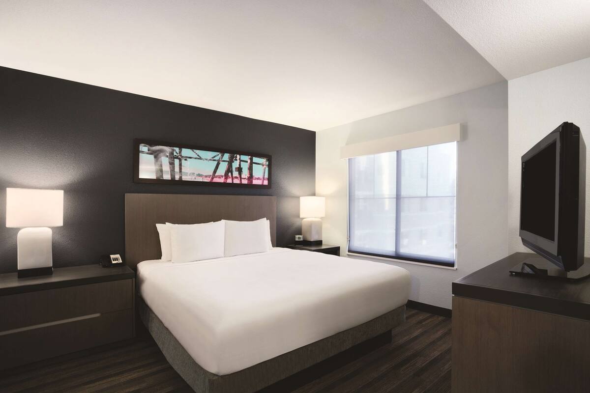 A hotel room with a large bed, two bedside tables with lamps, a flat-screen TV, a window with blinds, and a wall-mounted artwork.