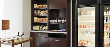 Una kitchenette con una mesa de madera, estantes llenos de bocadillos, un enfriador de vino y un refrigerador.