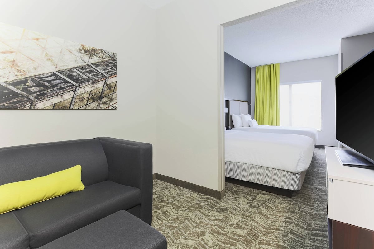 Een hotelkamer met een grijze bank, een bed met wit beddengoed, een televisie op een witte standaard, en een schilderij aan de muur.