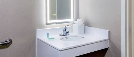 A bathroom with a white sink, a mirror with built-in lighting, a soap dispenser, and a tissue roll.
