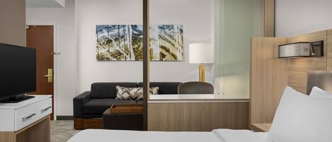 Ett hotellrum med en säng, ett skrivbord med en tv, en soffa, en lampa och två tavlor på väggen.