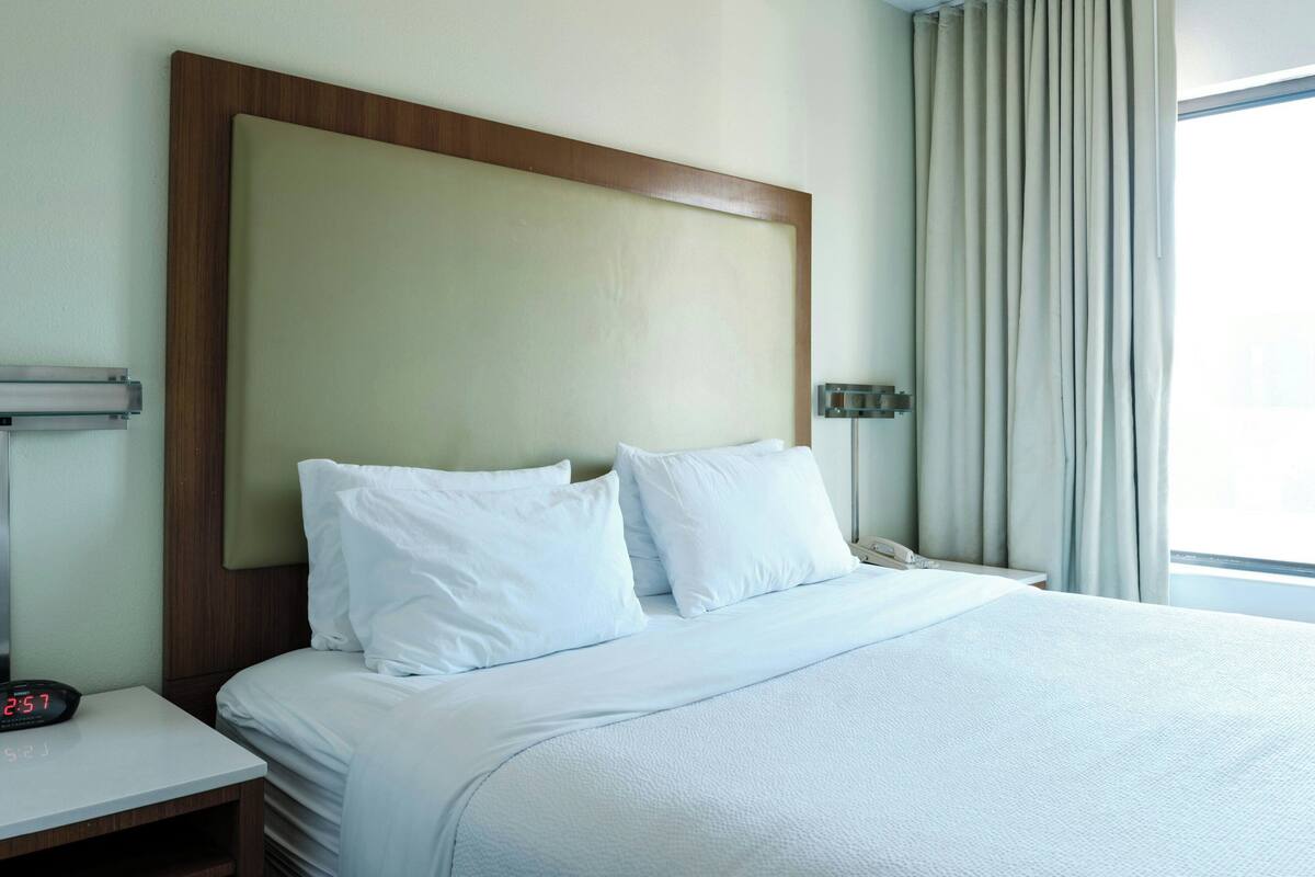 Habitación de hotel con cama, mesita de noche con reloj, y ventana con cortinas.