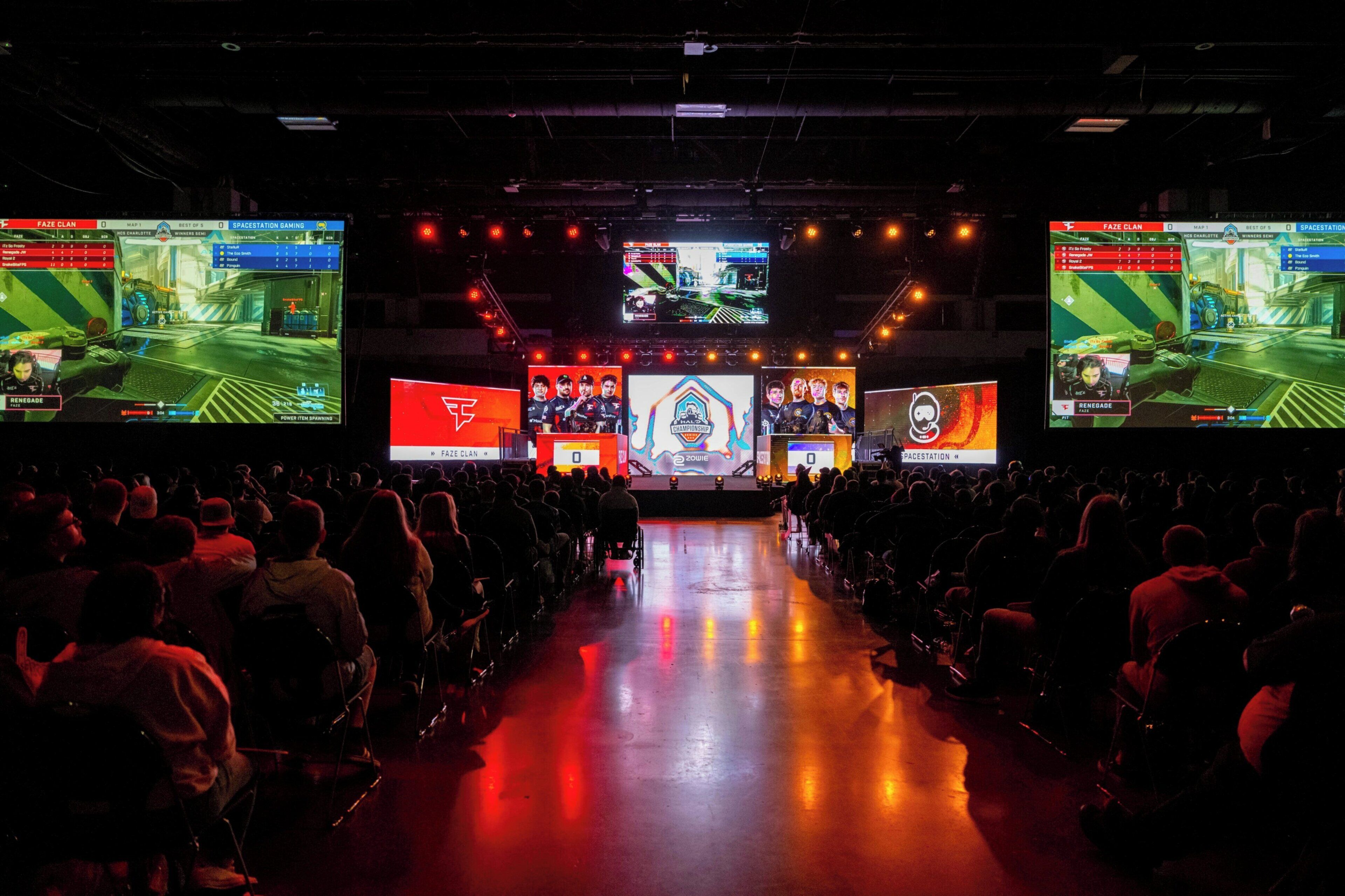 Un espacio interior amplio con varias pantallas mostrando contenido de videojuegos, un escenario central y un público sentado en filas.