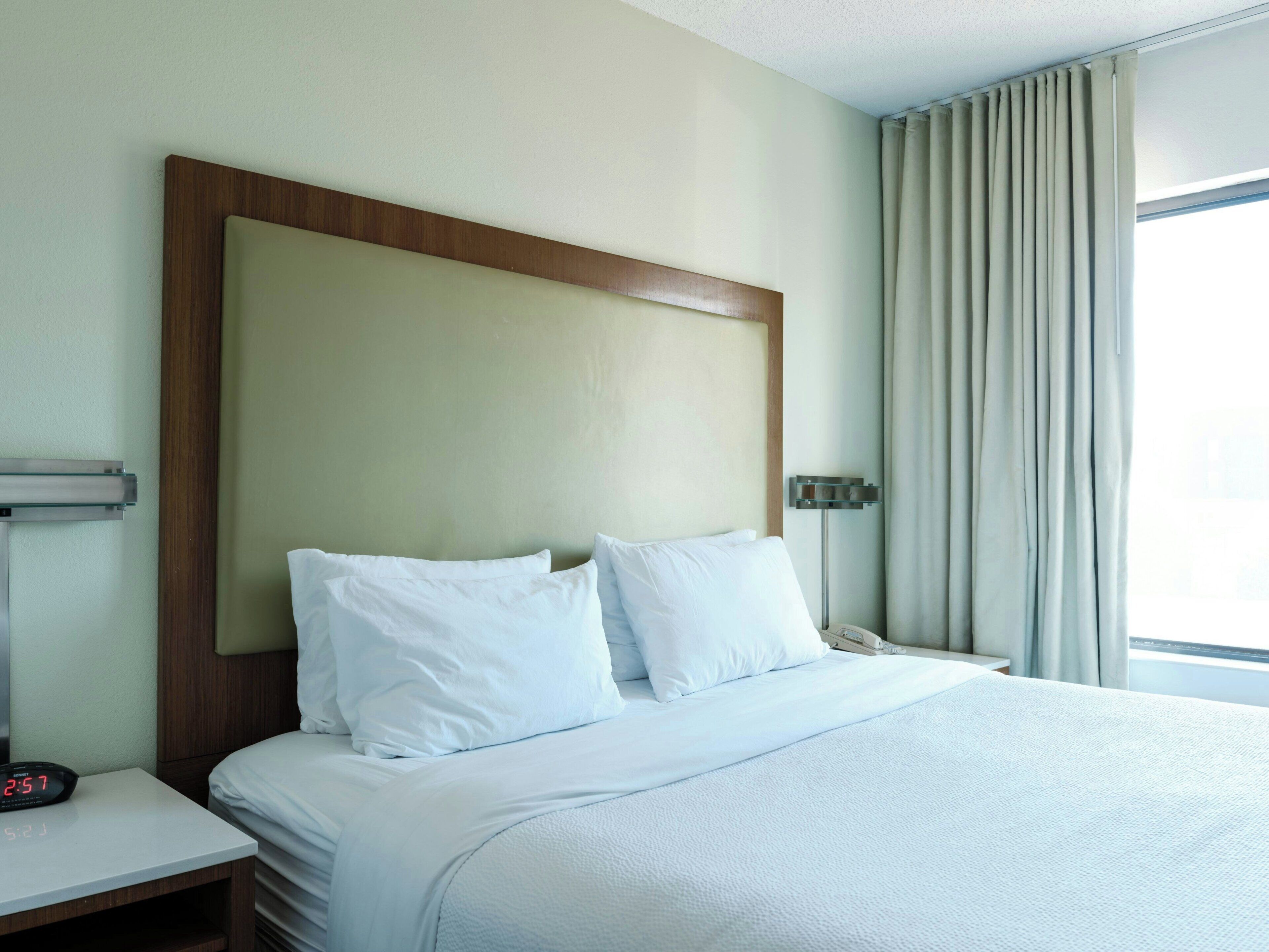 A hotel room with a bed, a nightstand with a clock, a bedside table, and a window with curtains.