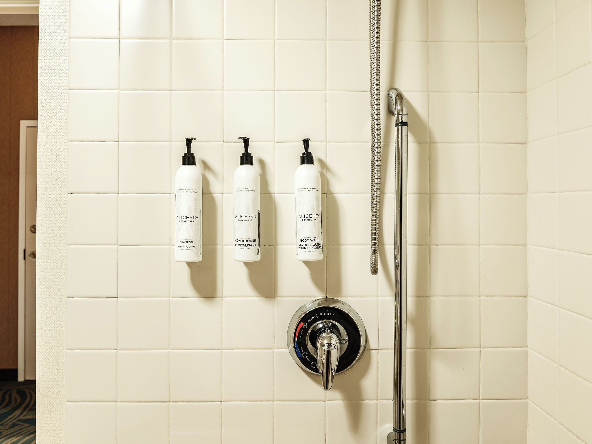 A bathroom with white tiles, a Kohler showerhead, and three bottles of Alice + Co Botanicals body wash.