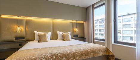 Una habitación de hotel moderna con una cama grande, un cabecero con iluminación integrada, vistas de la ciudad a través de amplias ventanas y una paleta de colores neutros.