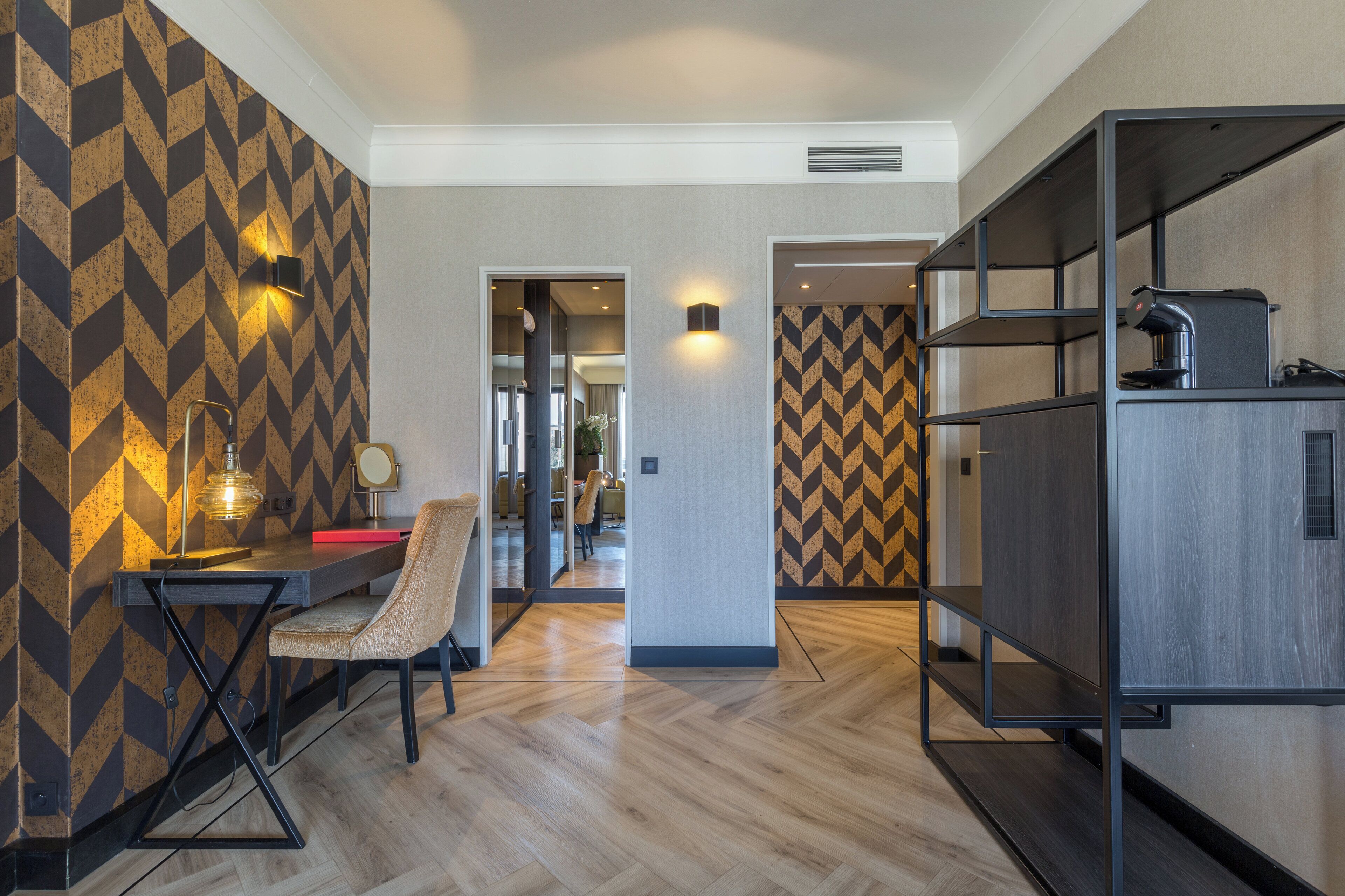 A modern hotel room with a wooden floor, a desk with a chair, and a built-in kitchen area.