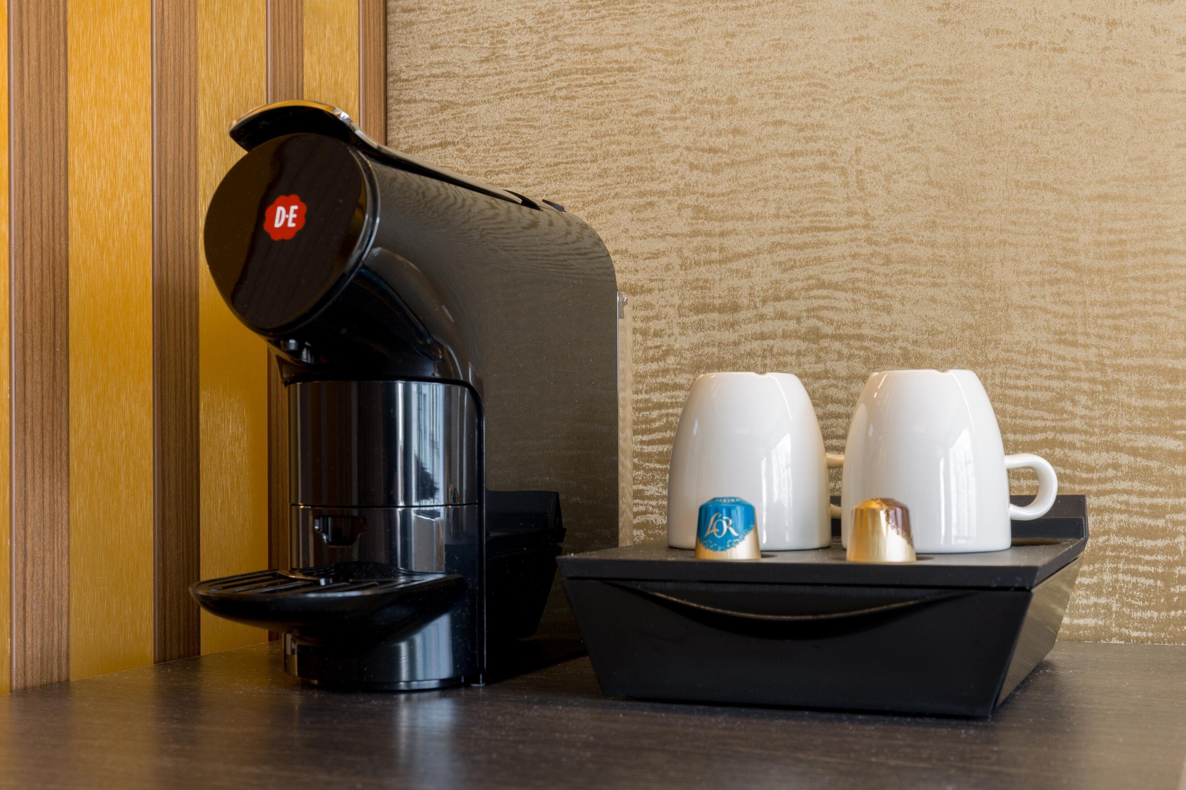 A coffee maker with a "DE" logo, two white cups, and a small container with a blue lid on a black tray.
