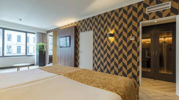 Een hotelkamer met twee bedden, een groot raam, een kamerplant en een behang met patroon.