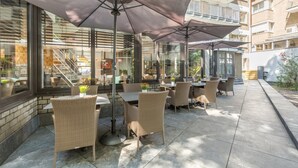Un patio al aire libre con mobiliario de mimbre y sombrillas grandes, con vistas a un edificio de varios pisos y balcones.