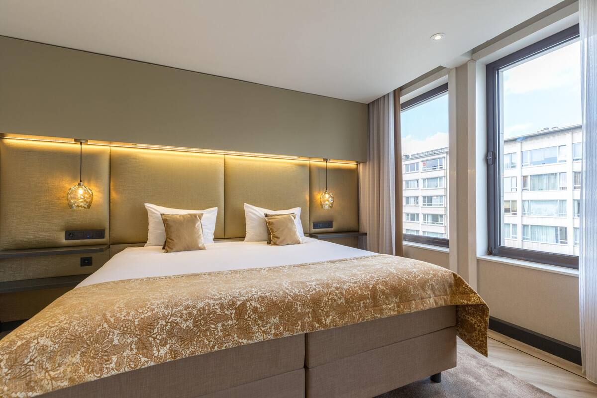 Une chambre d’hôtel moderne dotée d’un grand lit, d’une tête de lit avec éclairage intégré, d’une vue sur la ville grâce à de grandes fenêtres et d’une palette de couleurs neutres.
