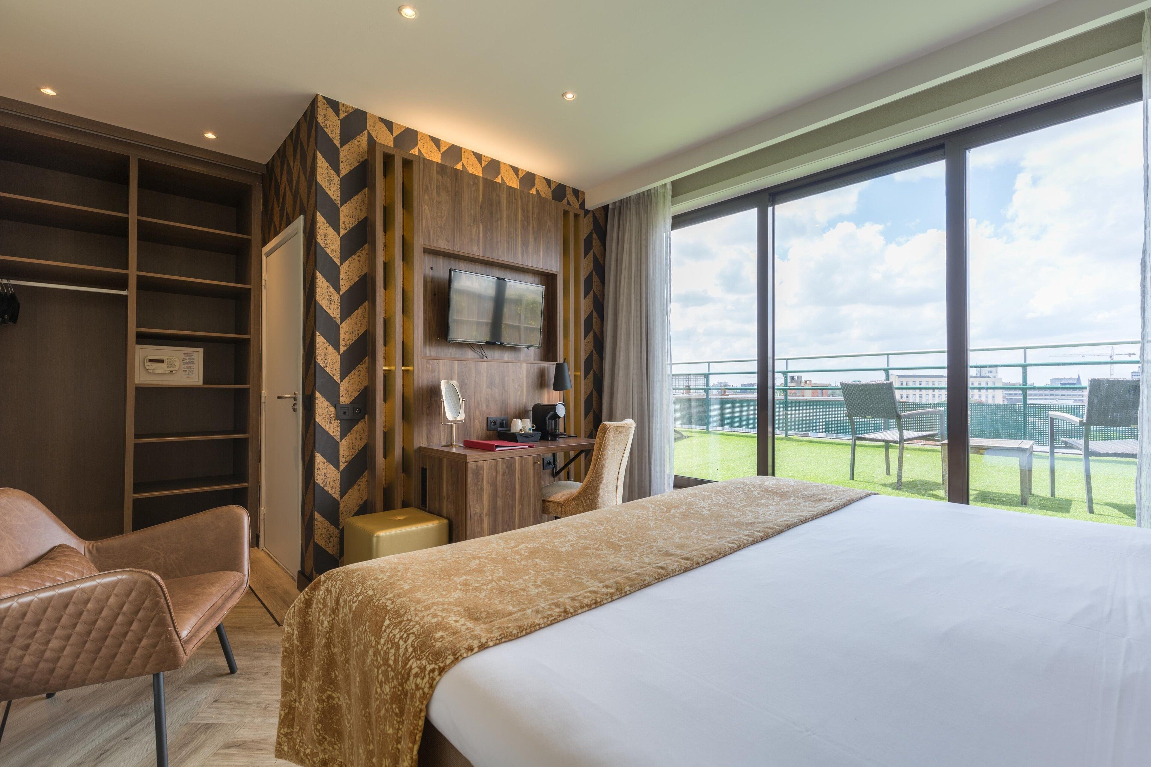 A hotel room with a large bed, a desk with a chair, a TV, and a balcony with outdoor seating.