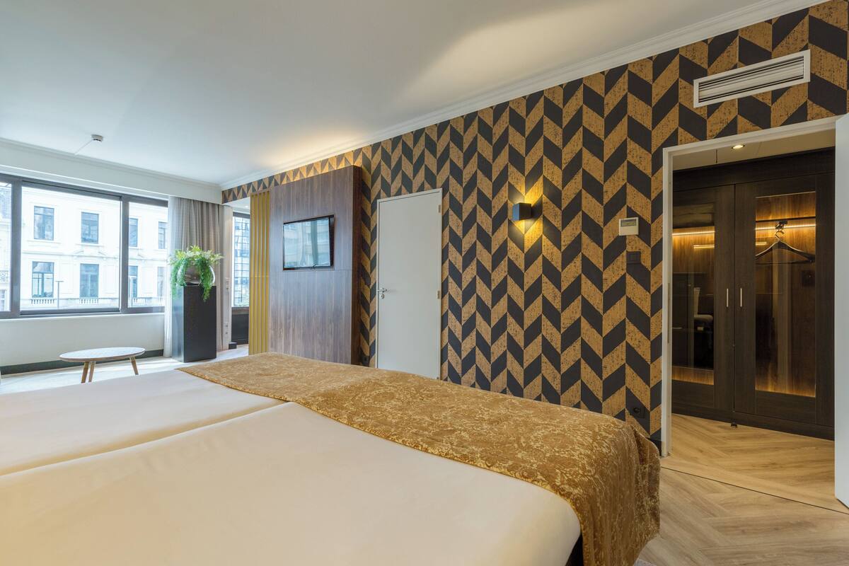 Une chambre d’hôtel avec deux lits, une grande fenêtre, une plante en pot et un mur à motifs.