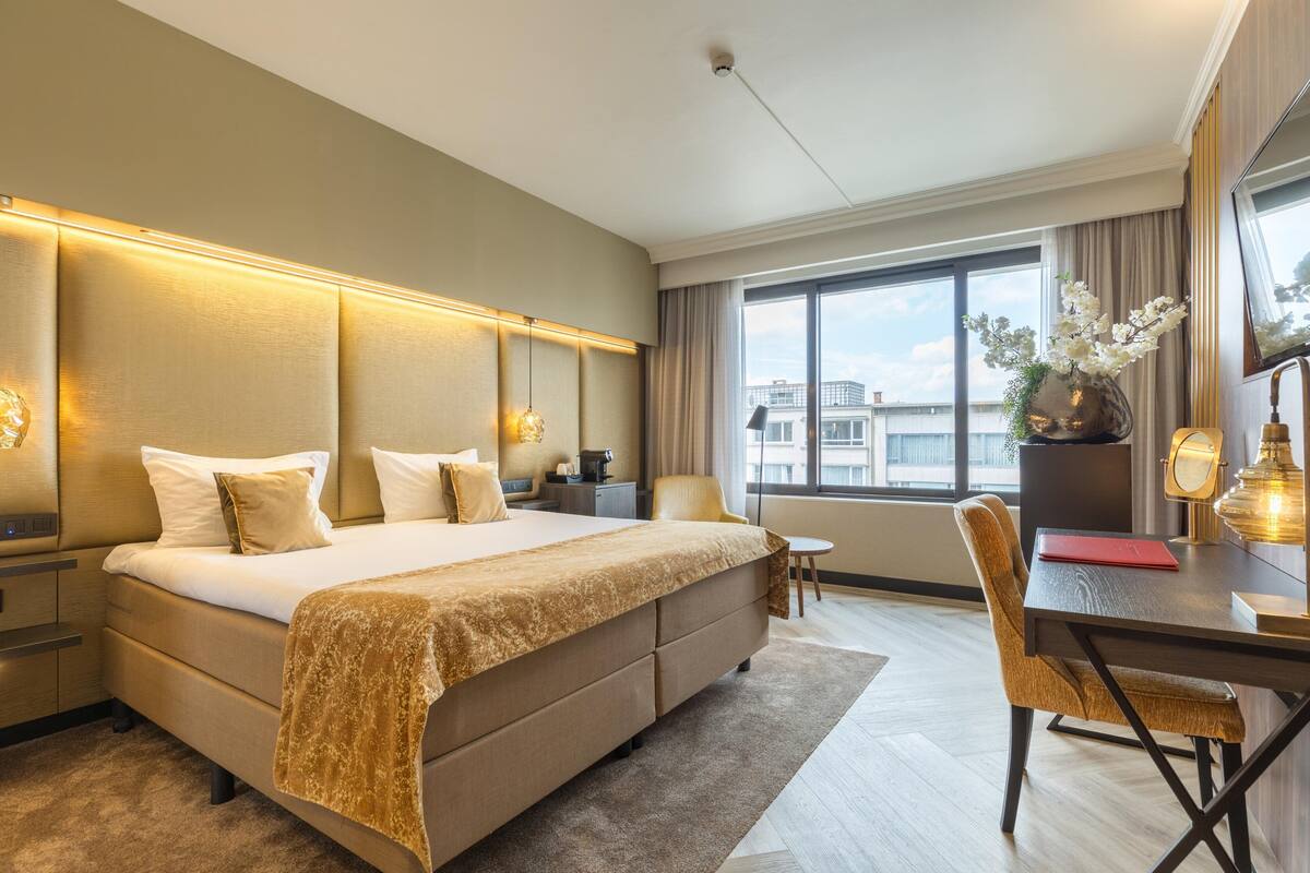 A hotel room with a large bed, a desk with a chair, a window with a view of buildings, and a vase with flowers on the desk.
