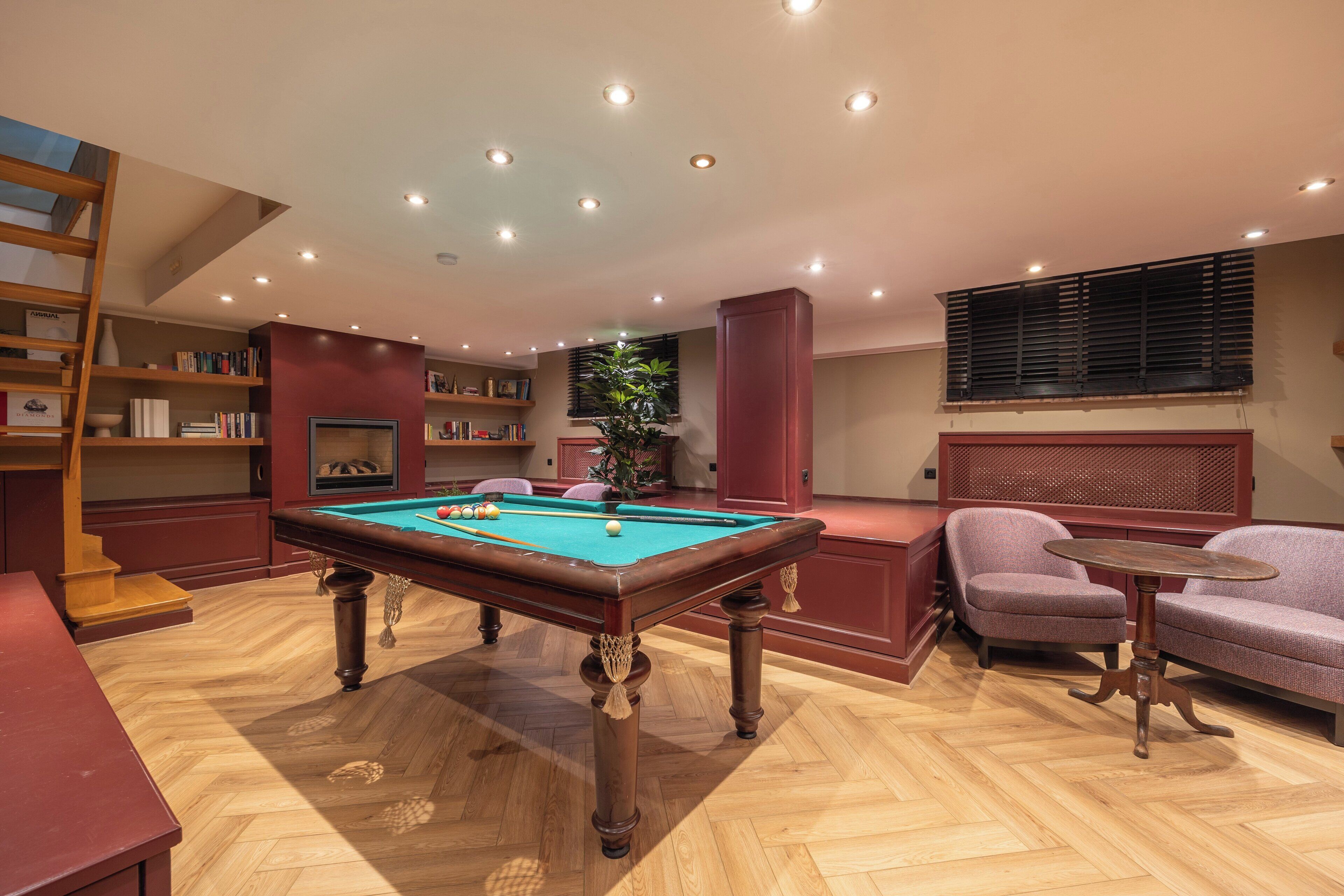Una mesa de billar en una habitación con piso de madera, una estantería, una silla y una ventana con persianas.