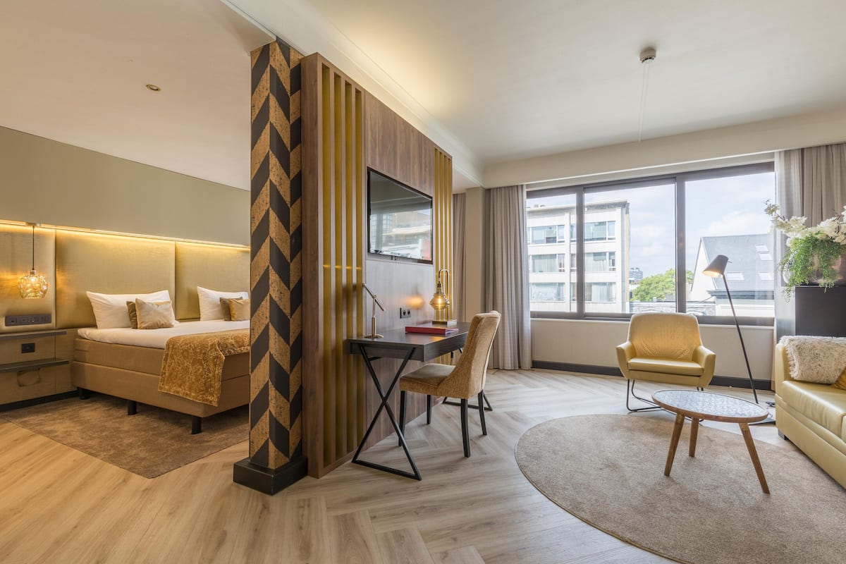 A modern hotel room with a large bed, a desk with a chair, a sofa, and a view of the city through large windows.