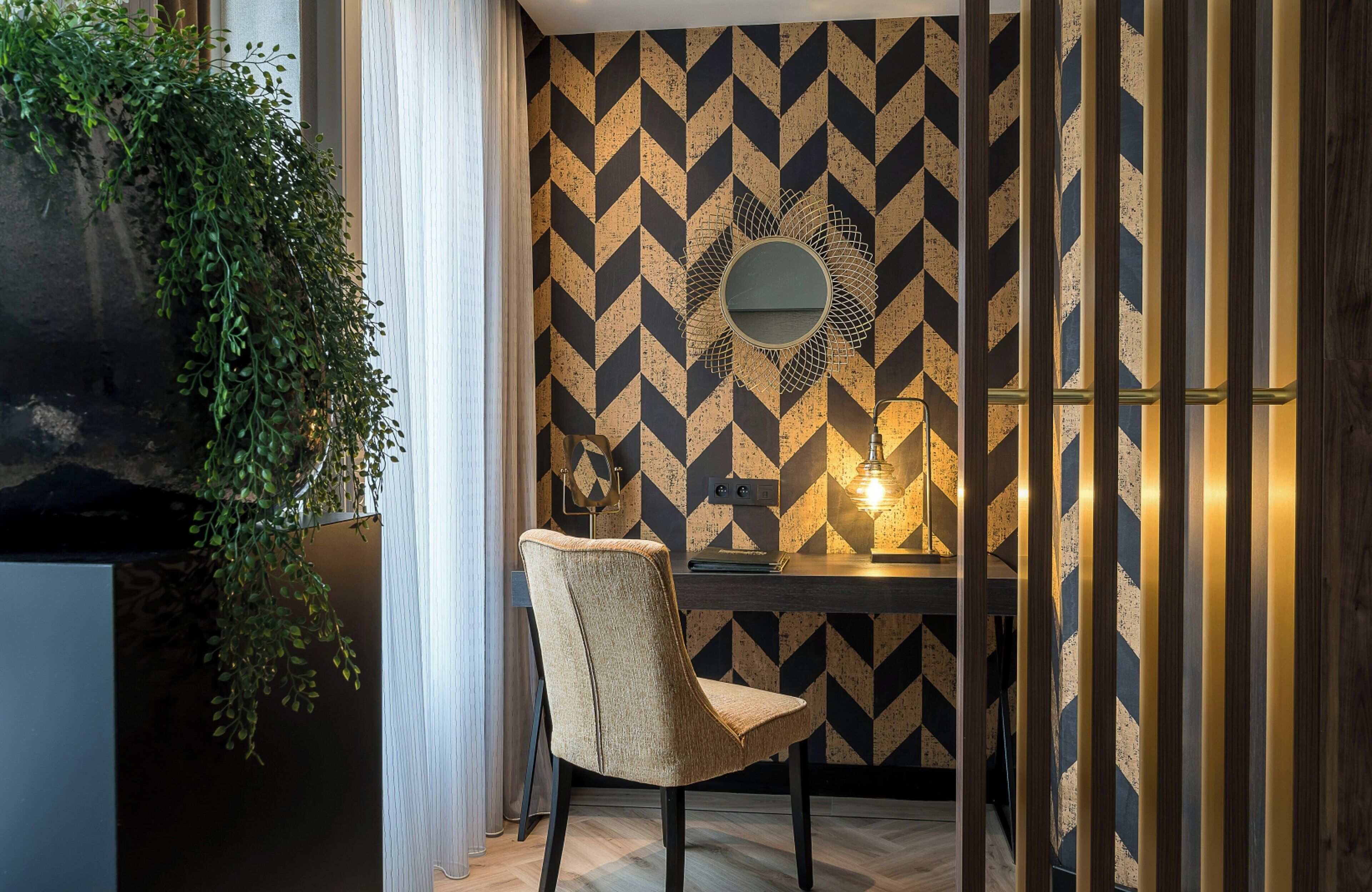 A room with a geometric patterned wall, a round mirror, a desk with a lamp, and a chair.
