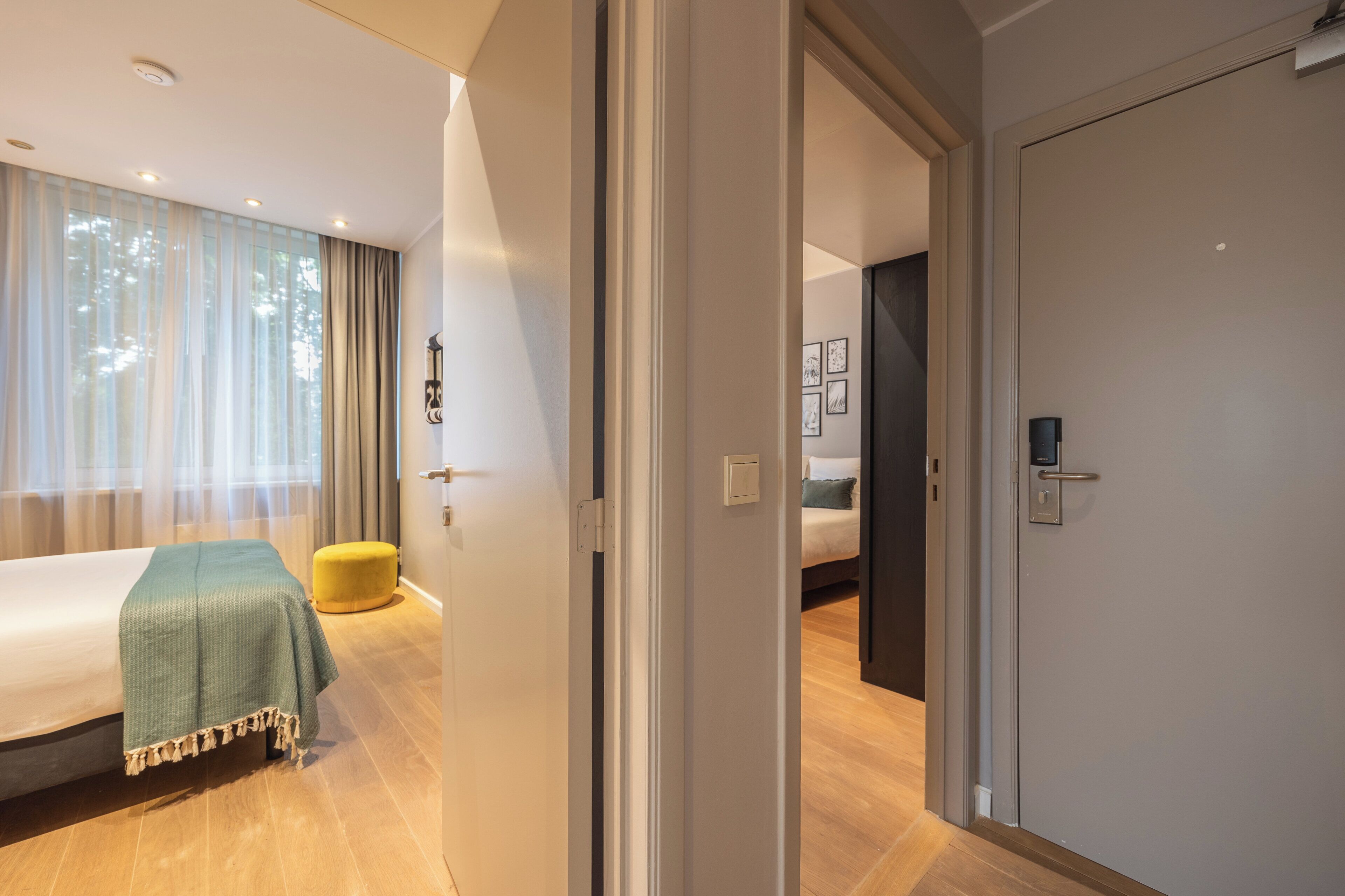 Ett modernt hotellrum med en säng, en liten gul pall och en dörr som leder till ett annat rum.