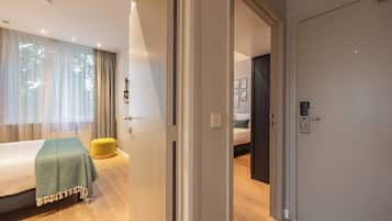 Ett modernt hotellrum med en säng, en liten gul pall och en dörr som leder till ett annat rum.
