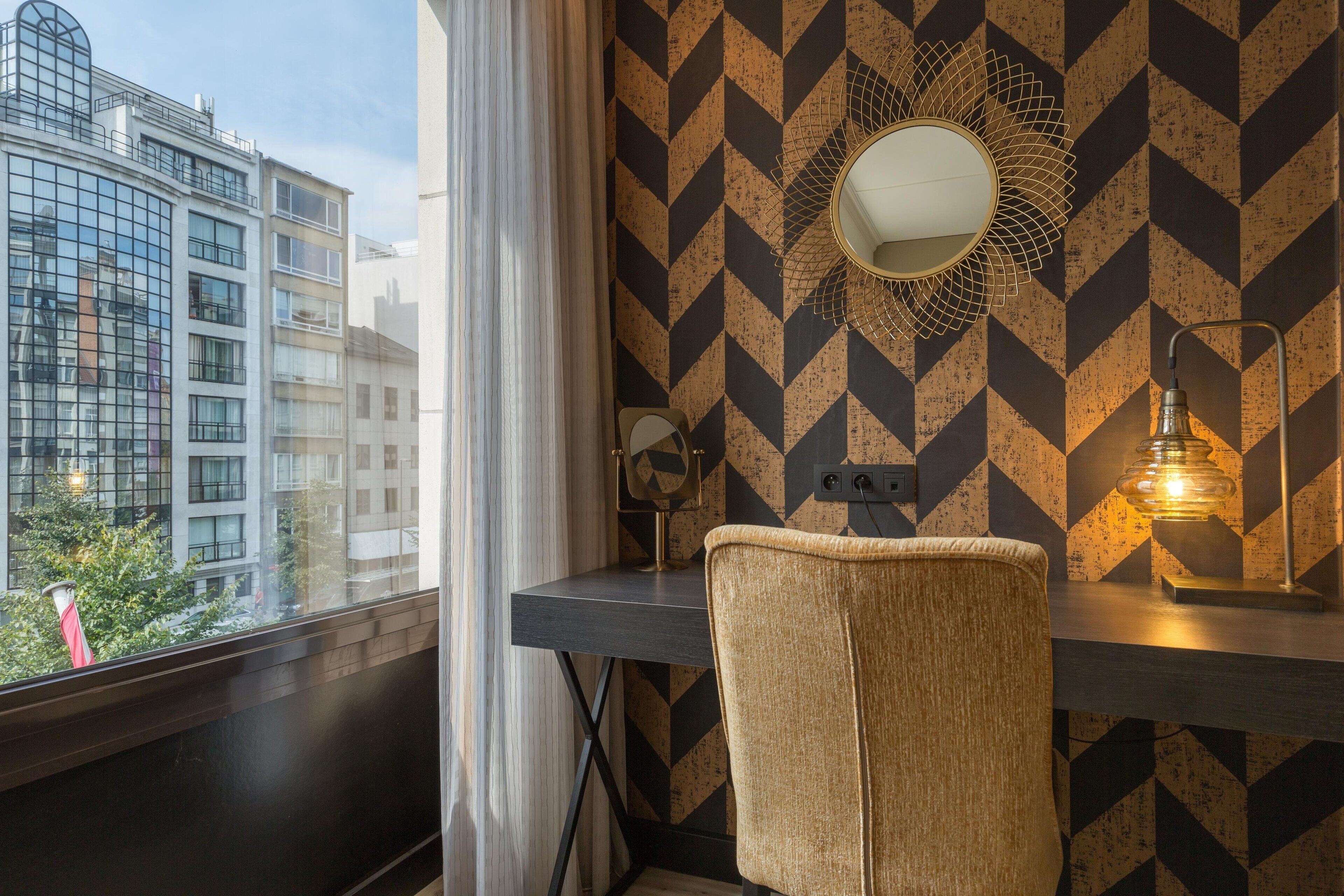 A modern hotel room with a geometric patterned wall, a round mirror, a desk with a lamp, and a view of buildings outside.