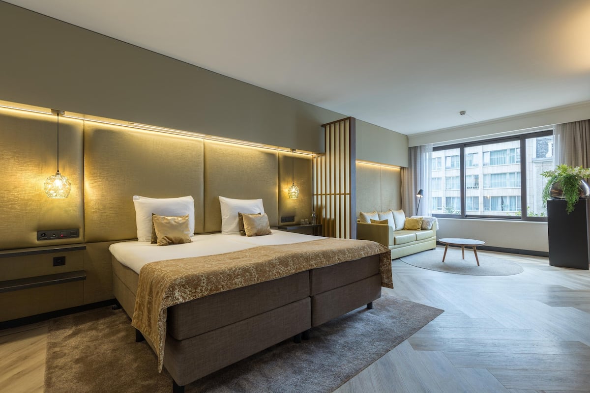A modern hotel room with a large bed, bedside tables, a sofa, a small table, and a large window.