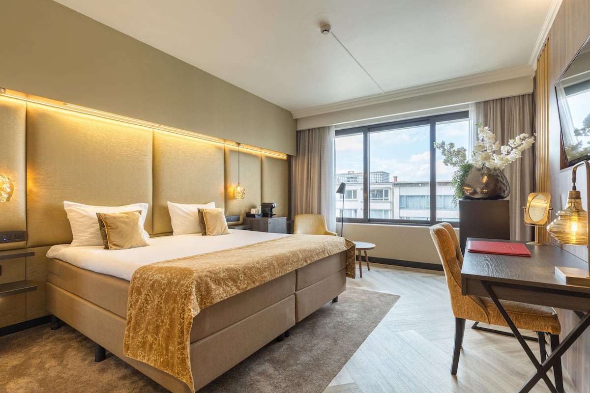 Une chambre d’hôtel avec un grand lit, un bureau avec une chaise, une fenêtre donnant sur des immeubles et un vase rempli de fleurs posé sur le bureau.