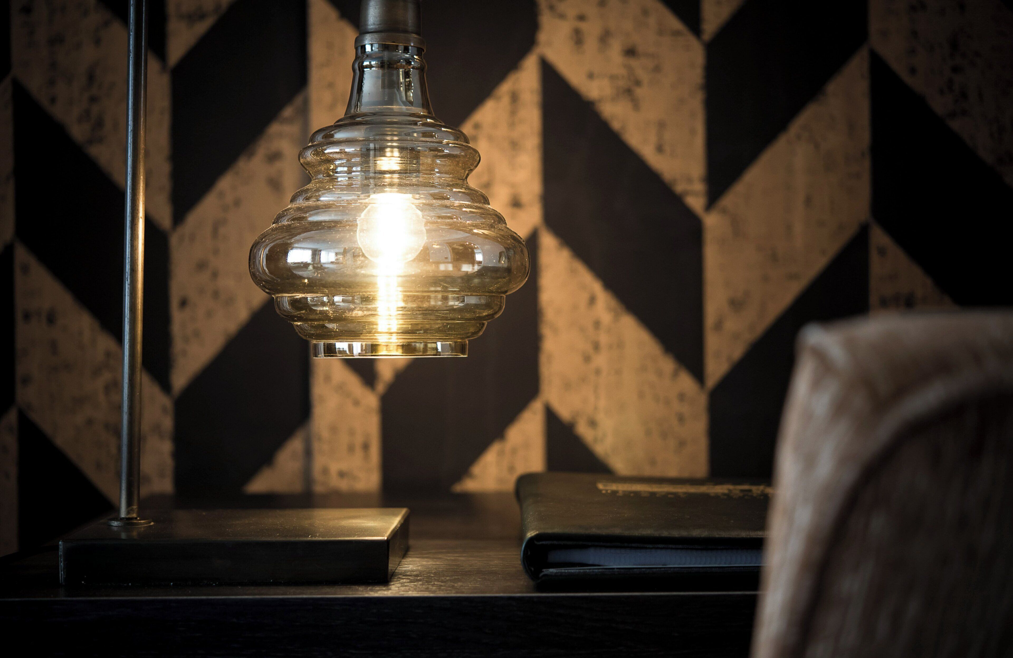 Uma luminária de mesa com cúpula de vidro transparente e base metálica, colocada sobre uma superfície de madeira, com uma parede ao fundo apresentando padrão geométrico.