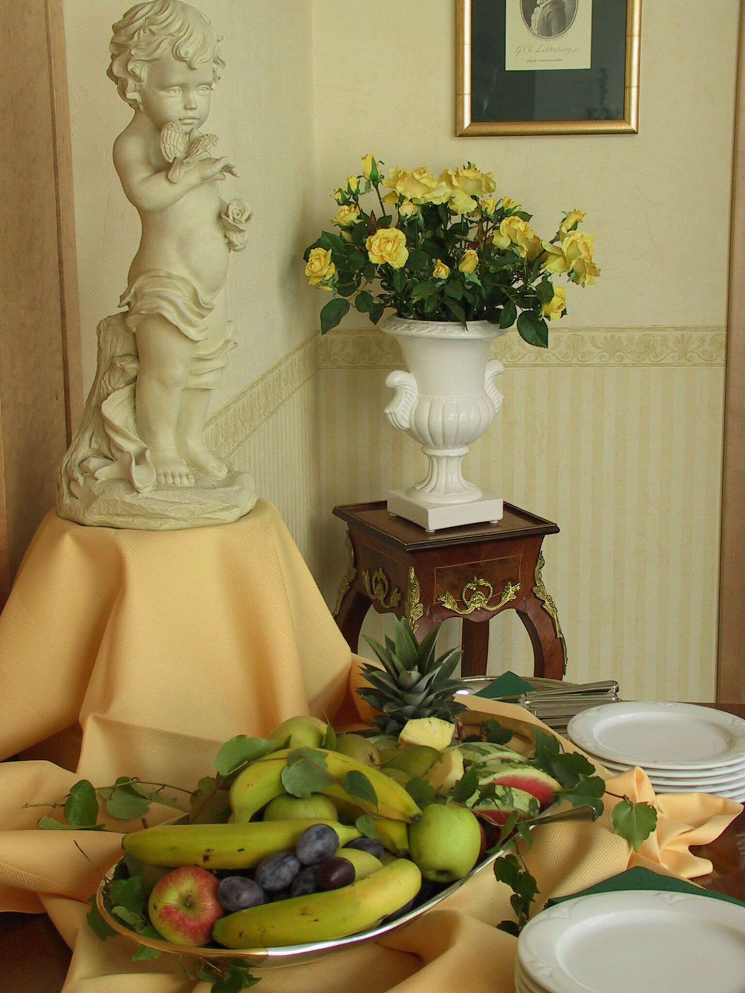 A table with a fruit bowl, a white vase with yellow flowers, and a statue.