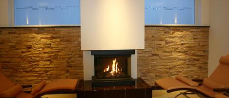 A fireplace with a stone wall backdrop, two windows with a view, and orange seating.