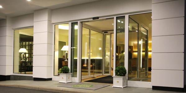 A hotel entrance with glass doors, a reception desk, and two potted plants.